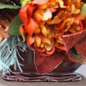 May include: A dark brown ceramic planter filled with an arrangement of orange and red flowers, green foliage, light blue sprigs, and a pale green pumpkin. The planter features a scalloped base and a glossy finish, accented by textured brown leaves.