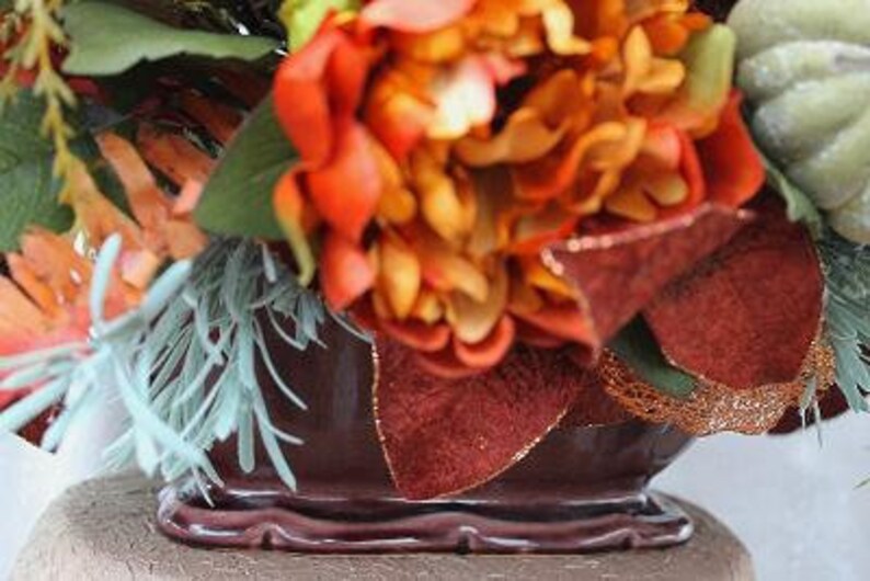 May include: A dark brown ceramic planter filled with an arrangement of orange and red flowers, green foliage, light blue sprigs, and a pale green pumpkin. The planter features a scalloped base and a glossy finish, accented by textured brown leaves.