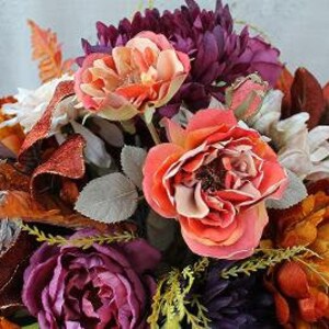 May include: A floral arrangement featuring artificial roses in shades of coral and orange, deep purple peonies, and white blossoms. The bouquet includes dark red and orange autumn-toned leaves, green foliage, and a decorative silver woven sphere.