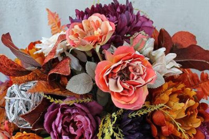 May include: A floral arrangement featuring artificial roses in shades of coral and orange, deep purple peonies, and white blossoms. The bouquet includes dark red and orange autumn-toned leaves, green foliage, and a decorative silver woven sphere.