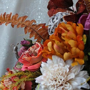 May include: A floral arrangement featuring a large white dahlia, vibrant orange peonies, and deep red glittered leaves. The bouquet includes a silver decorative orb, a dried brown fern frond, and small purple rosebuds, creating a textured display.