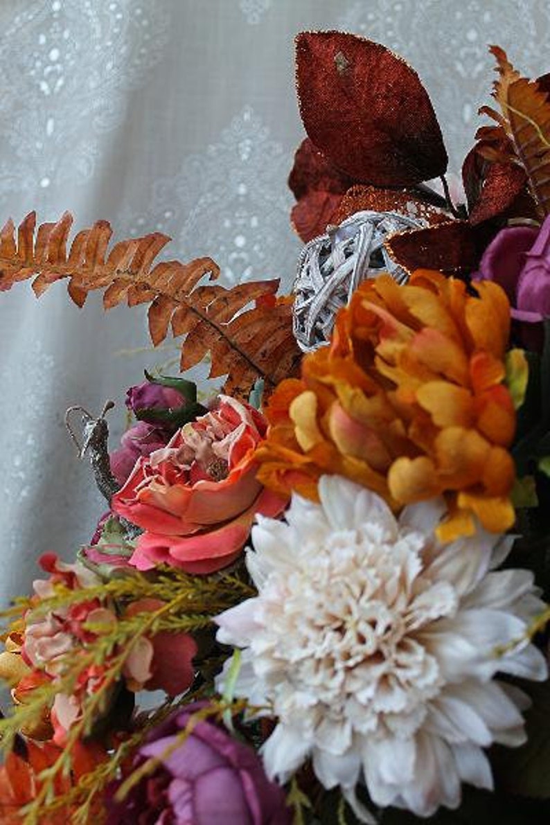 May include: A floral arrangement featuring a large white dahlia, vibrant orange peonies, and deep red glittered leaves. The bouquet includes a silver decorative orb, a dried brown fern frond, and small purple rosebuds, creating a textured display.