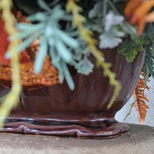May include: A dark brown ceramic bowl with a fluted design and a raised base, filled with an arrangement of artificial foliage including orange, green, and light blue sprigs, along with red glittered accents and textured greenery.