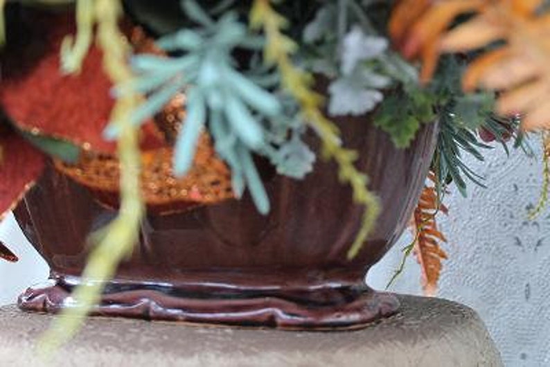 May include: A dark brown ceramic bowl with a fluted design and a raised base, filled with an arrangement of artificial foliage including orange, green, and light blue sprigs, along with red glittered accents and textured greenery.