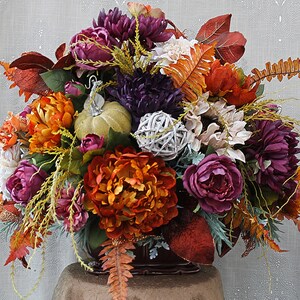 May include: A floral arrangement featuring deep purple, burnt orange, and cream-colored artificial flowers, including peonies and chrysanthemums. The display incorporates a small light green pumpkin, a white woven sphere, and various autumn-toned leaves.