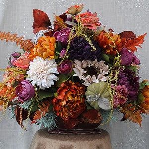 May include: A floral arrangement featuring artificial flowers in shades of deep purple, burnt orange, cream, and muted green. The bouquet includes peonies, chrysanthemums, sunflowers, and ferns, accented with a small fabric pumpkin in a dark container.