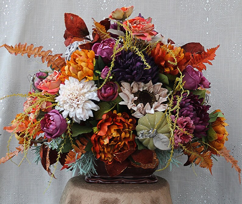 May include: A floral arrangement featuring artificial flowers in shades of deep purple, burnt orange, cream, and muted green. The bouquet includes peonies, chrysanthemums, sunflowers, and ferns, accented with a small fabric pumpkin in a dark container.