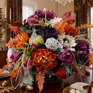 May include: A floral centerpiece featuring deep purple and vibrant orange peonies, a light green decorative pumpkin, a white woven sphere, and autumn-toned fern leaves. The arrangement is displayed in a dark container on a wooden table.