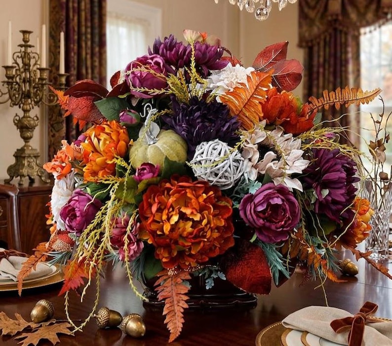 May include: A floral centerpiece featuring deep purple and vibrant orange peonies, a light green decorative pumpkin, a white woven sphere, and autumn-toned fern leaves. The arrangement is displayed in a dark container on a wooden table.