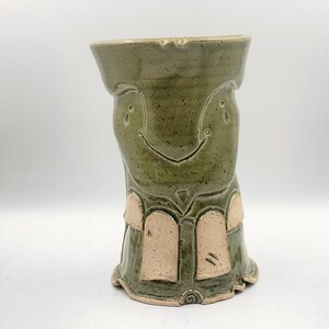 May include: Handmade ceramic vase in shades of green with a textured surface. The vase features a smiling face design and decorative arch shapes in a lighter color. The base has a unique, textured design.