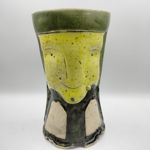 May include: A handmade ceramic vase with a face design. The vase features a yellow face with dark green accents and a dark green top. The base has a dark green and beige design. The vase is approximately 8 inches tall.