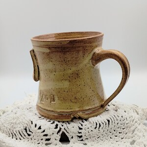 May include: A handmade ceramic mug with a brown and tan glaze. The mug has a unique shape with a handle and a small spout. The mug is sitting on a white crocheted doily. The mug has the number "201" written on the side.