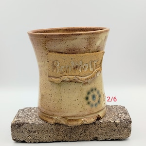 May include: A tan ceramic mug with a brown glaze and a white banner that reads "Red Wolf" in a handwritten font. The mug has a blue and white design on the side and the number "2/6" written on the side.