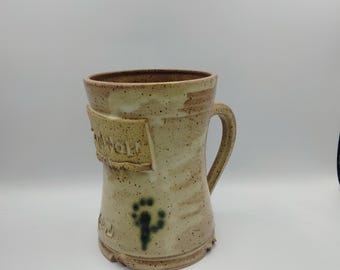 Puede incluir: Una taza de cerámica beige con asa marrón. La taza tiene un esmalte moteado marrón y blanco y un diseño floral verde y azul. La taza tiene la palabra "Wolf" escrita en ella.