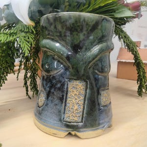 May include: A green and blue ceramic mug with a face design. The mug has a textured surface with small white dots and a brown rim.