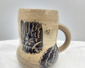 Puede incluir: Una taza de cerámica beige con asa marrón y diseños abstractos negros. La taza tiene un cuerpo redondeado y un borde ligeramente acampanado.