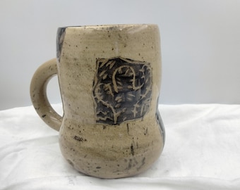Puede incluir: Una taza de cerámica con un esmalte moteado marrón y blanco. La taza tiene un asa y un diseño negro en la parte delantera.
