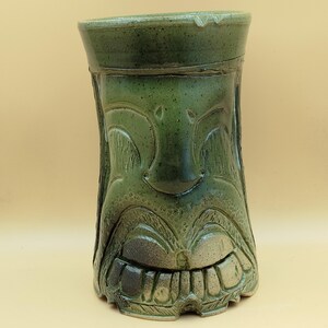 Può includere: Una tazza tiki in ceramica verde con un design a volto scolpito. La tazza presenta un volto sorridente con denti e lineamenti dettagliati. La tazza ha una forma cilindrica e una glassa verde scuro.