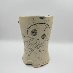 May include: A white ceramic mug with a black and white drawing of a skull with a dripping design. The mug has a speckled glaze and a blue rim.