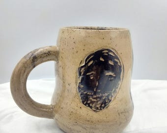 Puede incluir: Una taza de cerámica beige con un diseño de cara marrón. La taza tiene un esmalte moteado y una asa redondeada.