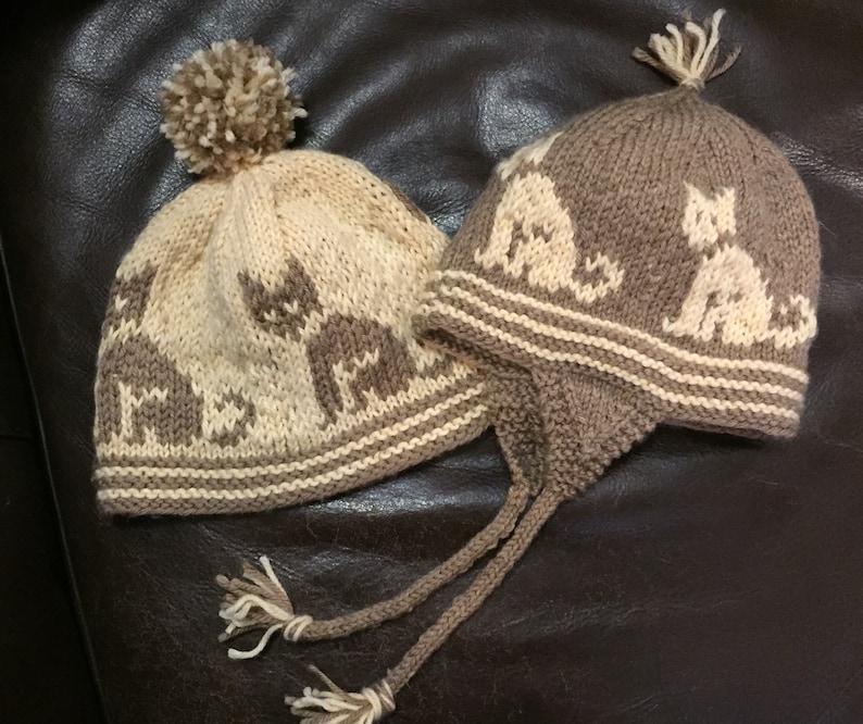 May include: Two knitted hats with a cat design. One hat is brown with a white cat design and the other is beige with a brown cat design. Both hats have a white pompom on top.