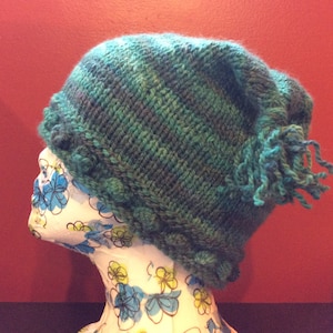 May include: A teal blue knitted hat with a fringe and a textured design. The hat is on a white mannequin head with a floral pattern.