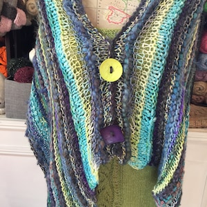 May include: A colorful striped knitted shawl with a large green button and a smaller purple button. The shawl is made with a variety of yarns and has a textured look.