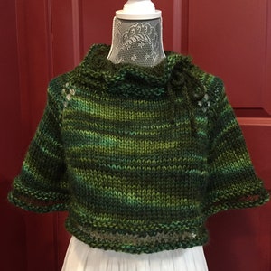 Könnte beinhalten: Ein grün-blauer gestrickter Pullover mit hohem Kragen und kurzen Ärmeln. Der Pullover hat einen Rüschenkragen und einen dekorativen Saum am unteren Rand.