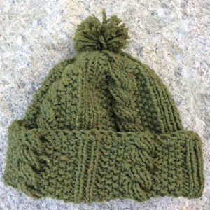 Op de afbeelding: Een groene gebreide muts met een textuurpatroon en een pompon bovenop. De muts heeft een omgeslagen rand en een kabelgebreid ontwerp. Het garen heeft spikkels van andere kleuren. De muts is ontworpen voor koud weer.