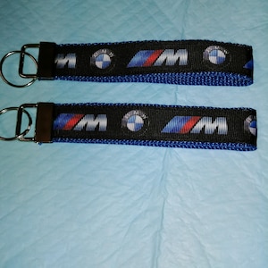 May include: Two black and blue keychains with a silver key ring. Each keychain has a repeating pattern of the BMW logo and the letters "M" in blue and red.