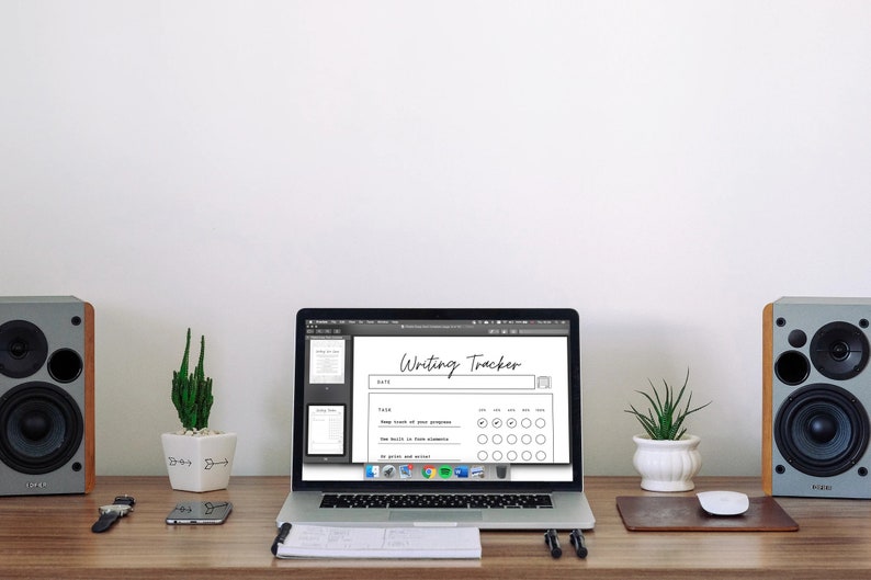 May include: A laptop displays a "Writing Tracker" document on a wooden desk. The desk also holds speakers, a watch, a phone, a mouse, and potted plants. The tracker has sections for dates and progress tracking. The background is a plain white wall.