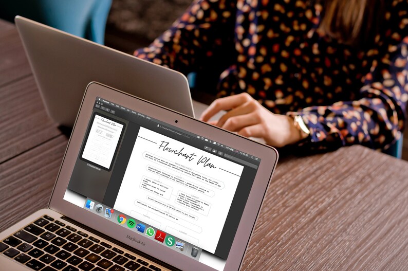 May include: Two MacBook Air laptops on a wooden table. The screen of the front laptop displays a "Flowchart Plan" document. The other laptop is open, and a person is working on it. The person is wearing a floral print top.
