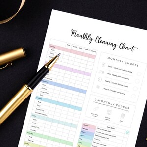 May include: A printed "Monthly Cleaning Chart" with a gold pen and ribbon on a black surface. The chart has a colorful grid for tracking chores. Gold safety pins are also visible.