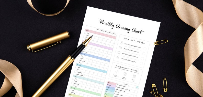 May include: A printed "Monthly Cleaning Chart" with a gold pen and ribbon on a black surface. The chart has a colorful grid for tracking chores. Gold safety pins are also visible.