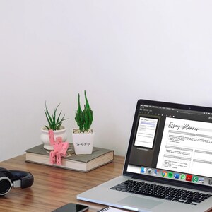 May include: A laptop displaying an "Essay Planner" document sits on a wooden desk, next to a pair of black headphones and a smartphone. Decorative plants and a pink sculpture add visual interest to the workspace.