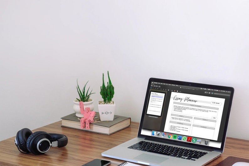 May include: A laptop displaying an "Essay Planner" document sits on a wooden desk, next to a pair of black headphones and a smartphone. Decorative plants and a pink sculpture add visual interest to the workspace.
