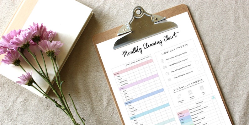 May include: A clipboard with a printed "Monthly Cleaning Chart" in black script. The chart has columns for weekly and monthly chores, with pastel color-coded sections. A stack of books and pink flowers are on the left.