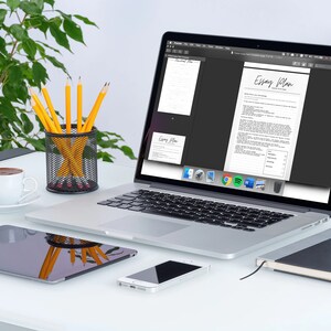 May include: A laptop displaying an "Essay Plan" document sits on a white desk, alongside a tablet, smartphone, and notebook. A cup of coffee, pencils in a wire mesh holder, and a green plant complete the workspace setup. The laptop screen shows the document in black and white.