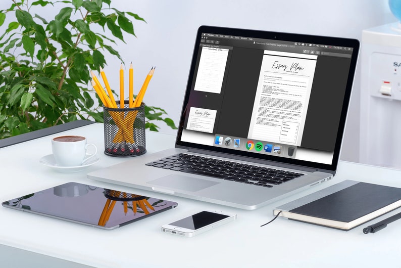 May include: A laptop displaying an "Essay Plan" document sits on a white desk, alongside a tablet, smartphone, and notebook. A cup of coffee, pencils in a wire mesh holder, and a green plant complete the workspace setup. The laptop screen shows the document in black and white.