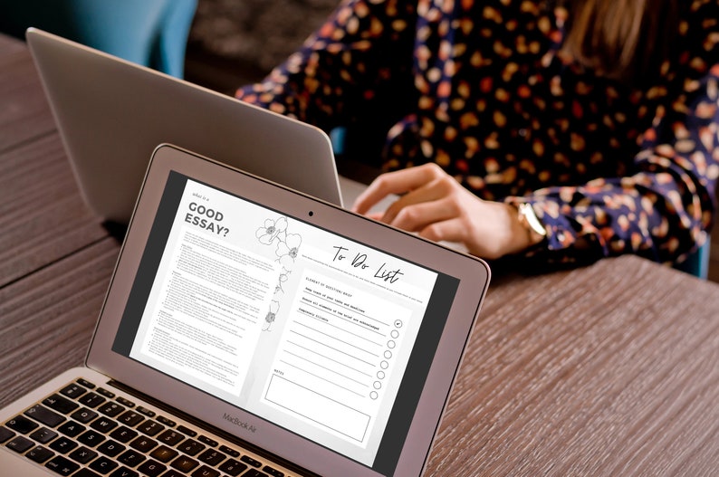 May include: Two open laptops on a wooden surface. One laptop displays the text "What is a GOOD ESSAY?" and a to-do list. The other laptop is partially visible. A person is working on the laptops.
