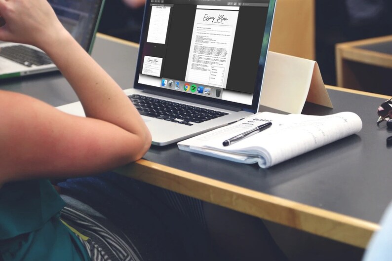 May include: A laptop displaying an "Essay Plan" document, with a notepad and pen on the desk. The laptop screen shows a document with text and a business card. The scene suggests a workspace or study environment.