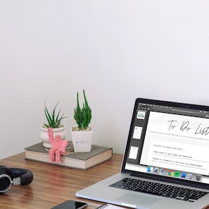May include: A laptop displaying a "To Do List" sits on a wooden desk, next to a pair of black headphones, a smartphone, and small potted plants. A pink decorative object and a book are also on the desk.