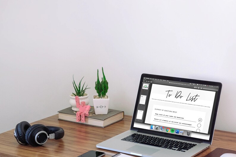 May include: A laptop displaying a "To Do List" sits on a wooden desk, next to a pair of black headphones, a smartphone, and small potted plants. A pink decorative object and a book are also on the desk.