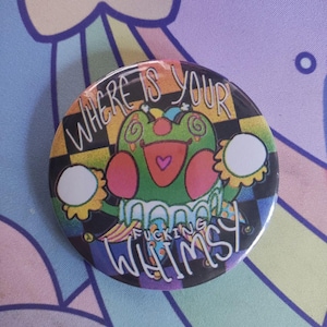 Pode incluir: Um botão redondo com um design de sapo de desenho animado colorido. O sapo é verde com detalhes em rosa e uma língua em forma de coração. O botão tem o texto "WHERE IS YOUR FUCKING WHIMSY" em branco e roxo.
