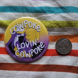 以下が含まれることがあります： 「cowpoke lovin cowpoke」という文字が書かれた、紫、黄、白のボタン。ボタンはストライプの布地に置かれている。