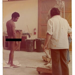 Puede incluir: Fotografía vintage de un estudio con una figura desnuda de pie sobre papel. Una persona con camisa blanca y pantalón oscuro trabaja en una escultura. Se ven dos grandes botes de basura y un extintor.