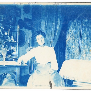 May include: A woman in a white blouse and a dark skirt sits in a chair in a room with blue patterned curtains. The image is a cyanotype, a photographic printing process that produces a blue-toned image.