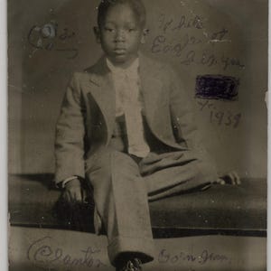 May include: Vintage black and white photograph of a young person seated, wearing a light-colored suit and dress shoes. The image has handwritten text and dates, including "1939" and "Born Jan. 1932". The photograph is aged and shows signs of wear.