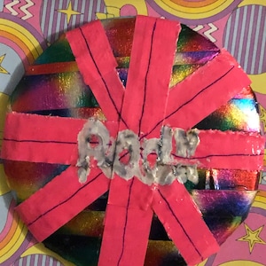 Könnte beinhalten: Eine runde, schillernde, regenbogenfarbene Scheibe mit rosa Klebeband, das in einem Sternenausbruch um sie herumgewickelt ist. Das Wort "Rock" ist in weißer Farbe auf die Scheibe geschrieben.