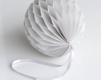 May include: A white, spherical paper decoration with a honeycomb design. The paper ball has a series of folded, fan-like sections that create a textured surface. A thin, white ribbon is attached for hanging.